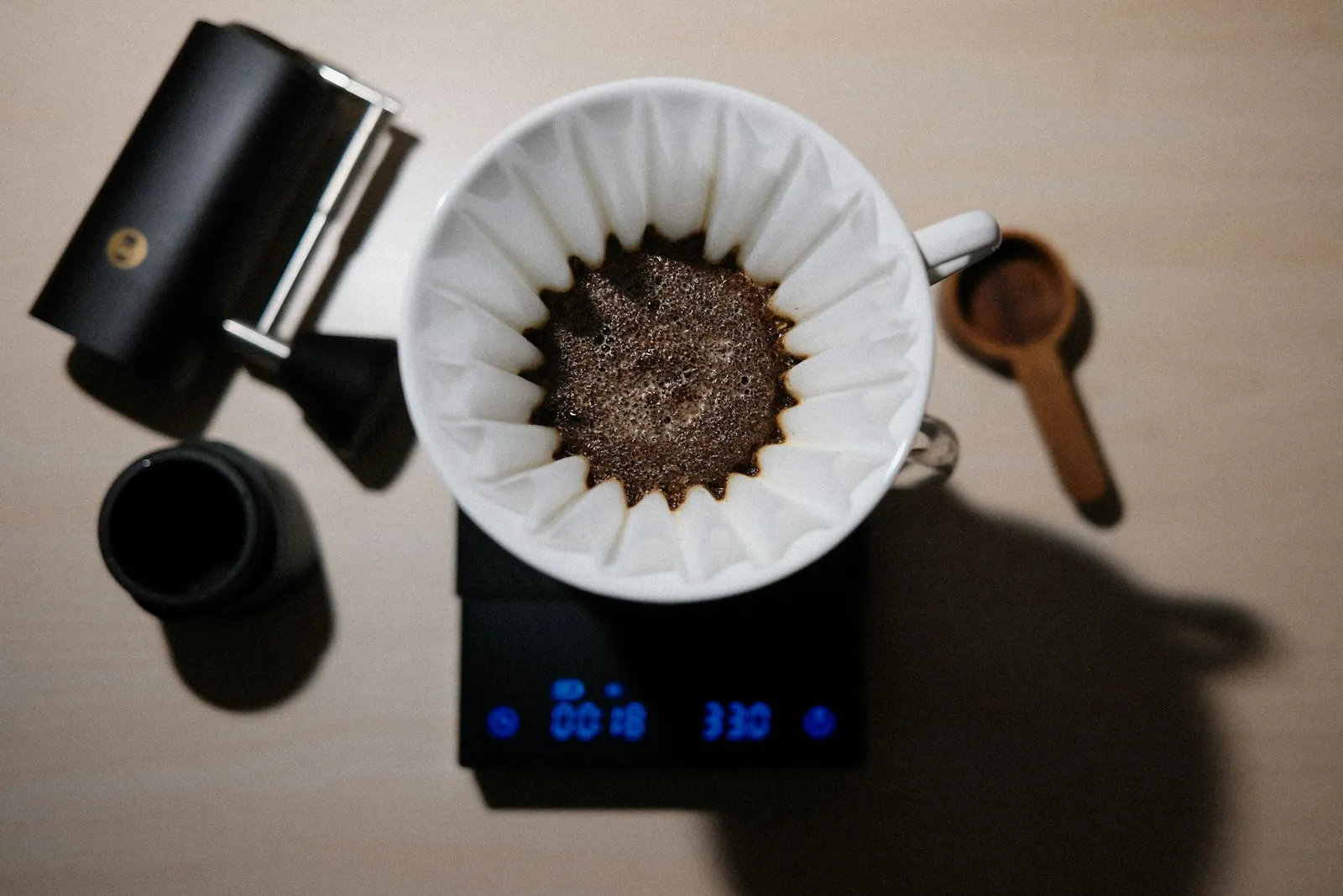 Coffee brewing in a white paper filter on a black digital scale, seen from above, beside a hand grinder and a wooden scoop